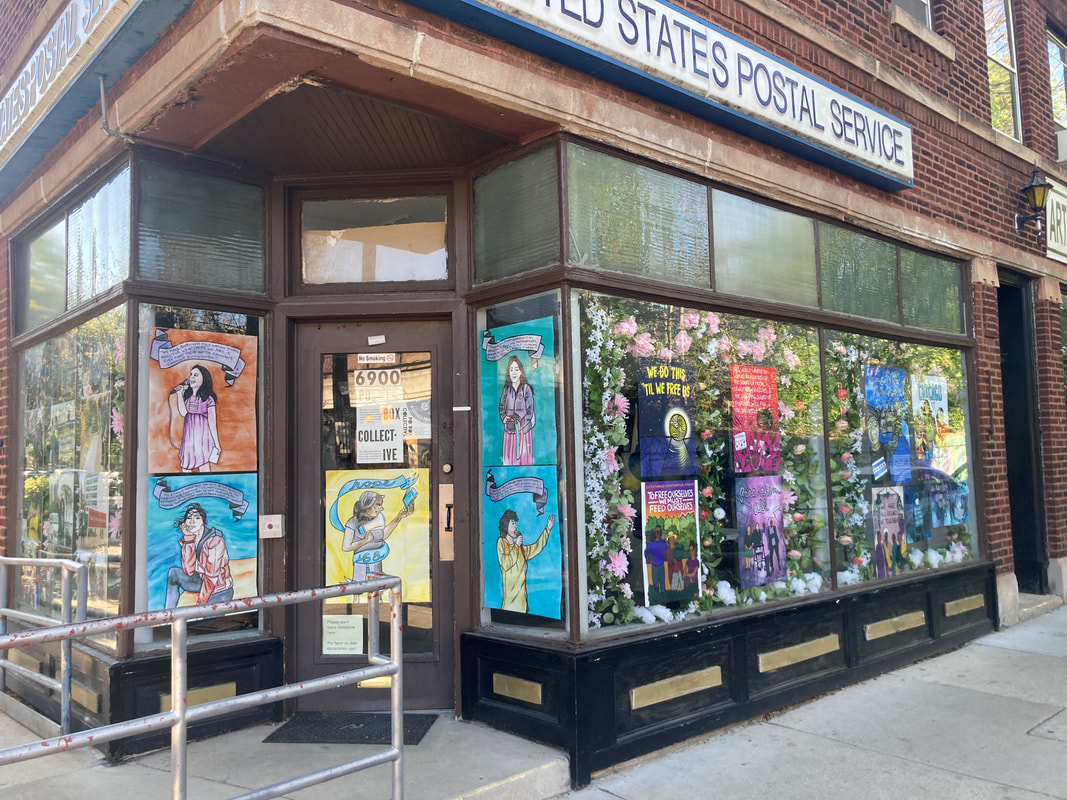 A photo of an old P.O. Box storefront transformed into an art space. My artwork is hanging in the windows with fake flower garland hanging in-between the artwork to create a frame. The entrance is wheelchair accessible and has a ramp going up.
